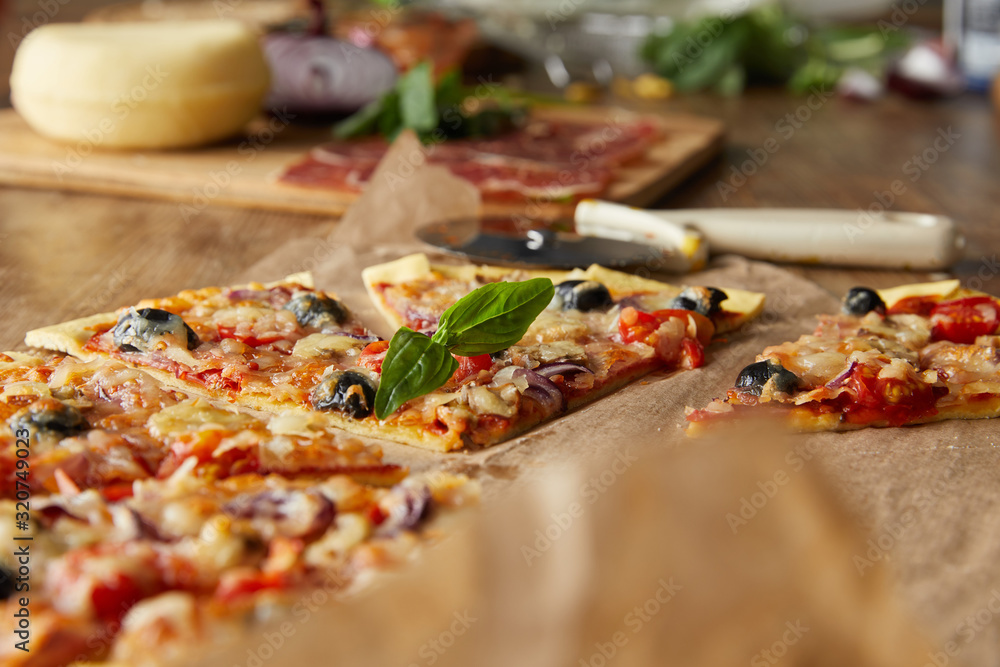 selective focus of cut delicious italian pizza with olives on baking paper near ingredients and pizza knife on wooden table selective focus of cut delicious italian pizza with olives on baking paper near ingredients and pizza knife on wooden table