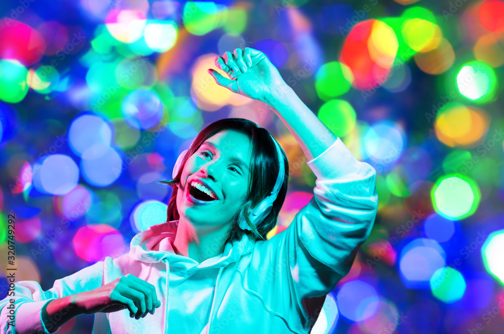 nightlife, technology and people concept - happy young woman in headphones wearing hoodie listening to music and dancing over night lights background nightlife, technology and people concept - happy young woman in headphones wearing hoodie listening to music and dancing over night lights background