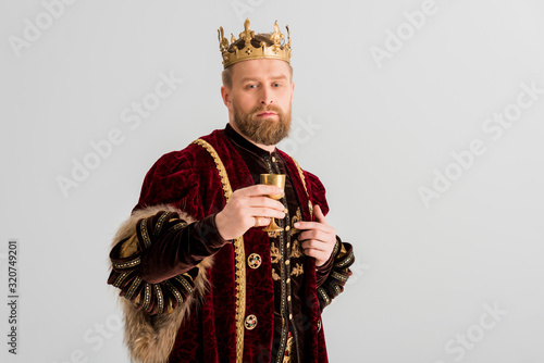 handsome king with crown holding cup and looking at camera isolated on grey