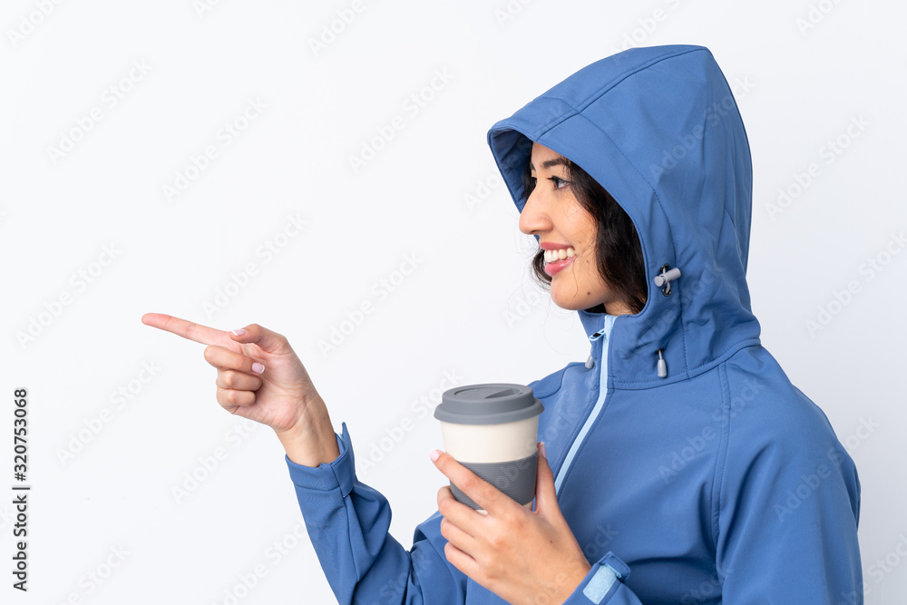 Mixed race woman wearing winter clothes with the hood on and holding a hot takeaway coffee over isolated white background pointing to the side to present a product Mixed race woman wearing winter clothes with the hood on and holding a hot takeaway coffee over isolated white background pointing to the side to present a product