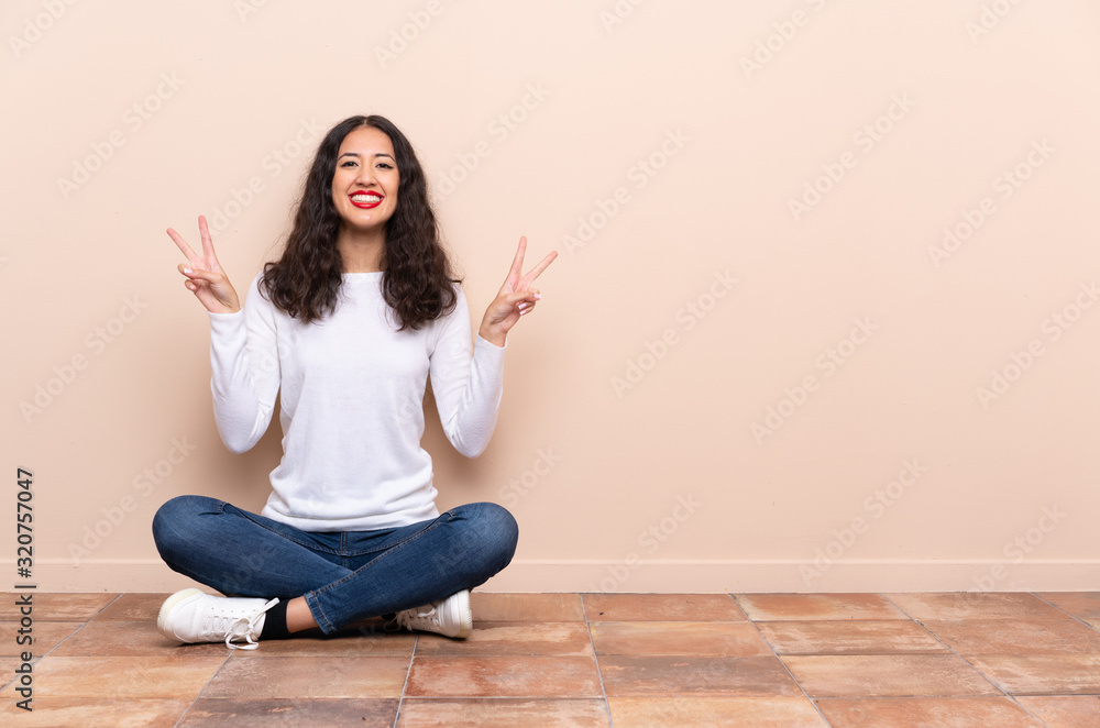 Young woman sitting on the floor showing victory sign with both hands Young woman sitting on the floor showing victory sign with both hands