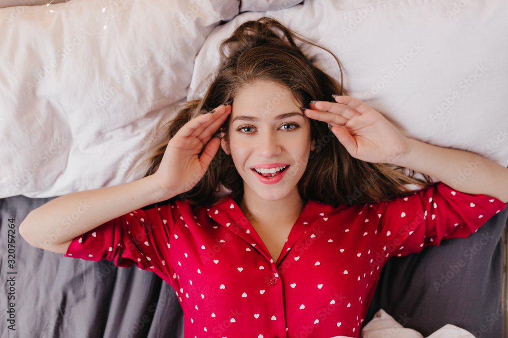 Fototapeta premium Amazing dark-haired female model enjoying morning in weekend. Indoor overhead photo of enthusiastic brunette girl lying in bed and laughing.