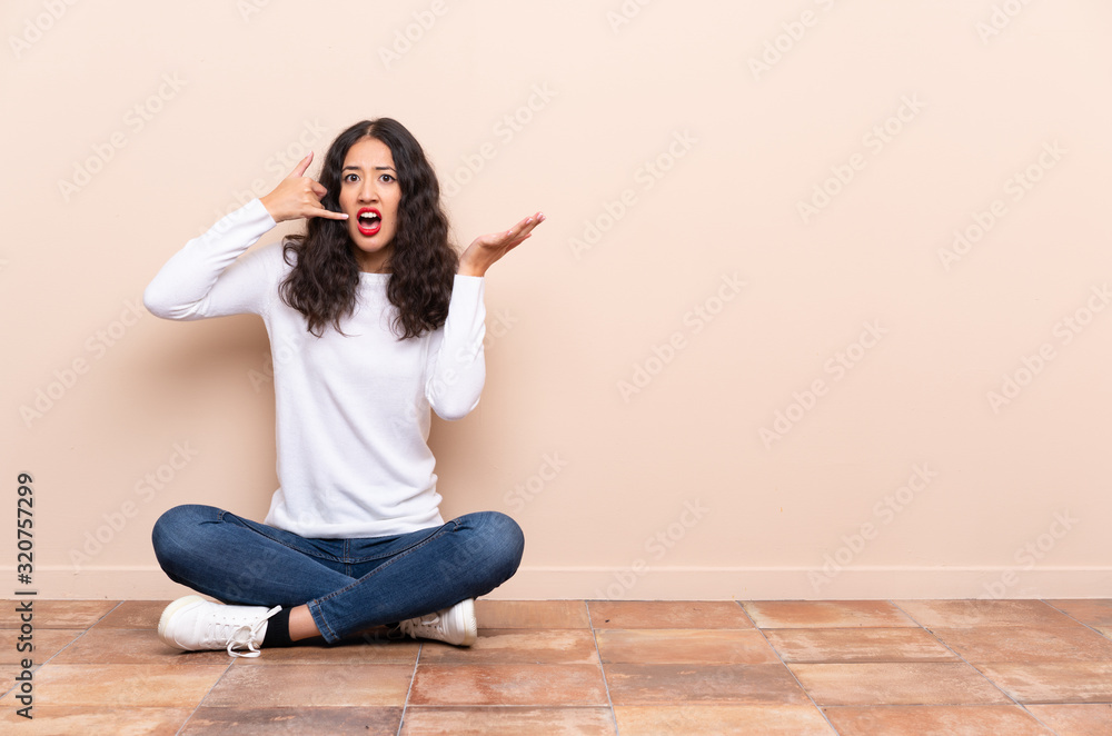Young woman sitting on the floor making phone gesture and doubting Young woman sitting on the floor making phone gesture and doubting
