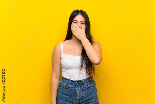 Young teenager Asian girl over isolated yellow background covering mouth with hands