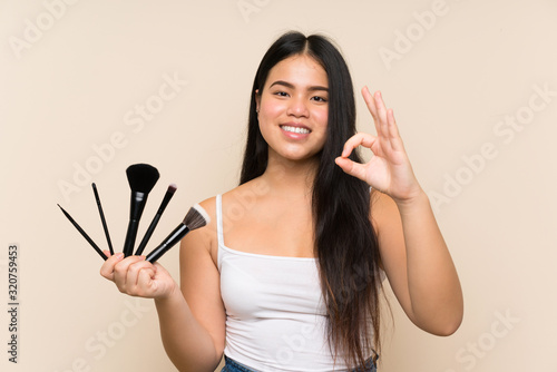 Young teenager Asian girl holding a lot of makeup brush showing ok sign with fingers
