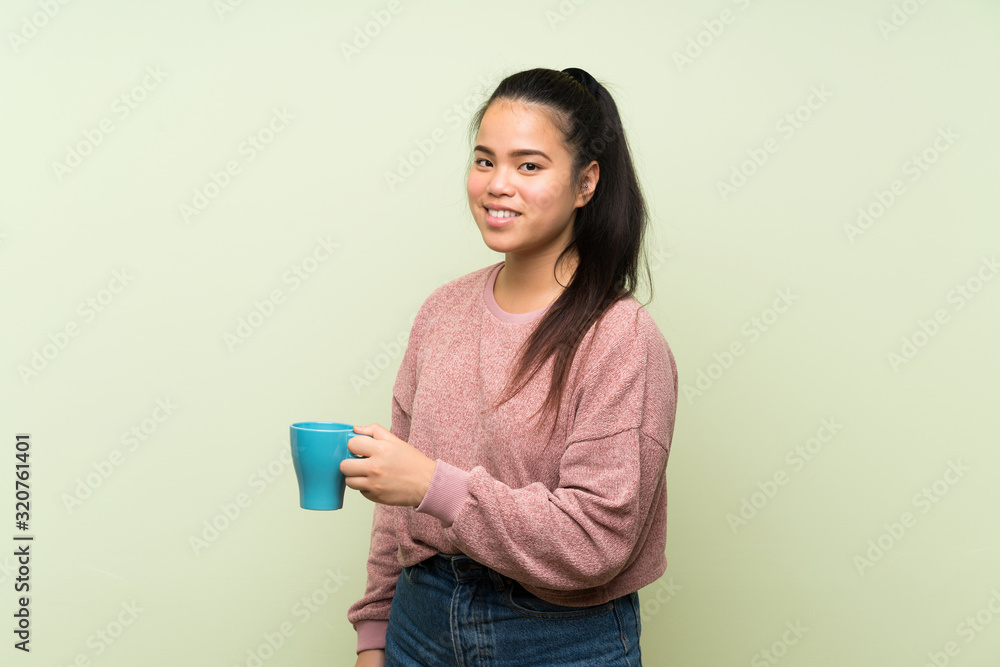 Young teenager Asian girl over isolated green background holding hot cup of coffee Young teenager Asian girl over isolated green background holding hot cup of coffee