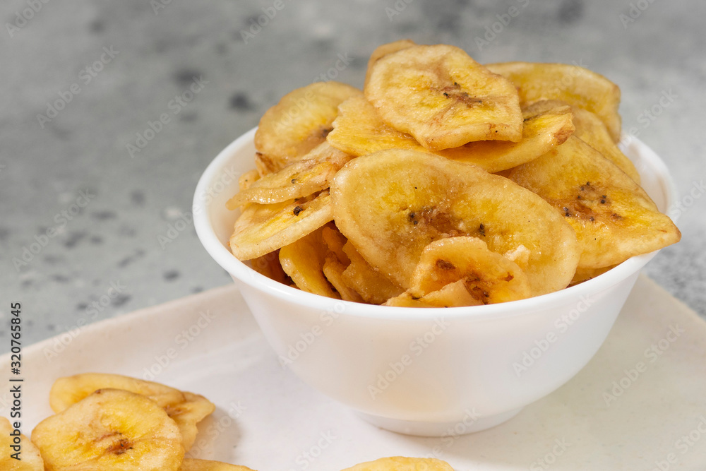 Dried bananas in a white ceramic bowl on a gray kitchen table. Banana chips. Vegetarian snack for proper nutrition Dried bananas in a white ceramic bowl on a gray kitchen table. Banana chips. Vegetarian snack for proper nutrition