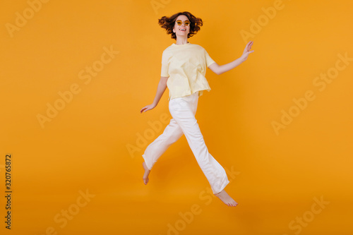 Full-length portrait of glad girl in yellow t-shirt and white pants running on orange background. Studio shot of wonderful caucasian woman dancing with pleasure.
