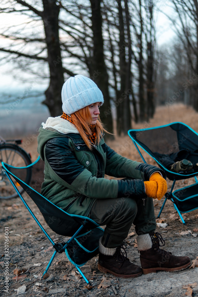 Girl sitting alone on light chairs by the fire. Cold autumn evening in the forest. The concept of an active lifestyle, travel, vacation. campfire, vertical photo Girl sitting alone on light chairs by the fire. Cold autumn evening in the forest. The concept of an active lifestyle, travel, vacation. campfire, vertical photo