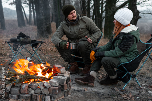 A couple of people are sitting on light chairs by the fire. Cold autumn evening in the forest. active lifestyle, travel, vacation. campfire. have a conversation over a cup of tea, dialogue © Sergey