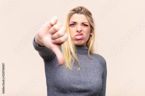 young pretty blonde woman feeling cross, angry, annoyed, disappointed or displeased, showing thumbs down with a serious look against flat wall