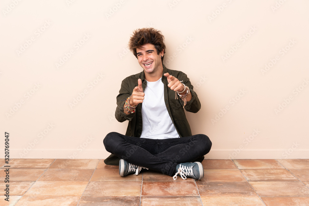 Young caucasian man sitting on the floor pointing to the front and smiling Young caucasian man sitting on the floor pointing to the front and smiling