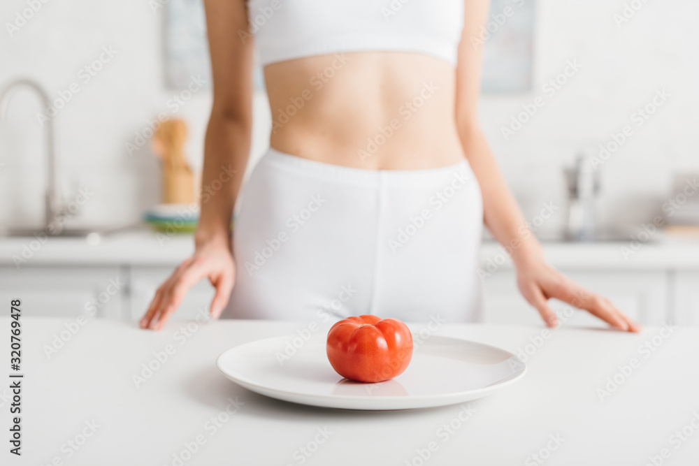 Selective focus of ripe tomato on plate and slim sportswoman near kitchen table