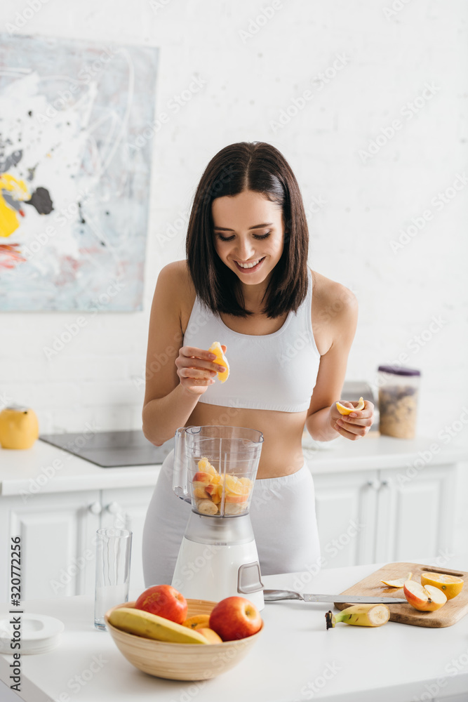 Smiling fit sportswoman adding fresh fruits in blender on kitchen table Smiling fit sportswoman adding fresh fruits in blender on kitchen table