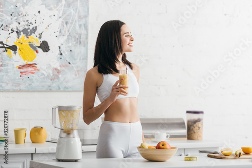 Bild auf Leinwand Attractive fit sportswoman smiling and holding glass of smoothie near measuring