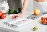 Selective focus of woman writing calories while weighing apple near fresh vegetables on kitchen table, calorie counting diet