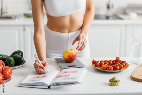 Cropped view of fit sportswoman writing calories while weighing apple on kitchen table, calorie counting diet photo