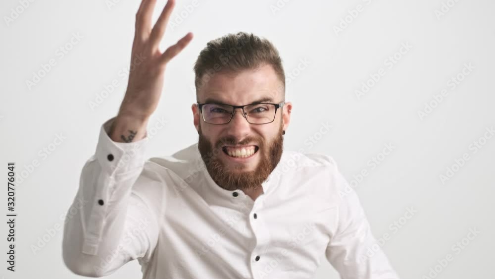 An attractive happy young bearded man wearing white shirt and glasses is gesturing while screaming to the camera isolated over white wall background An attractive happy young bearded man wearing white shirt and glasses is gesturing while screaming to the camera isolated over white wall background