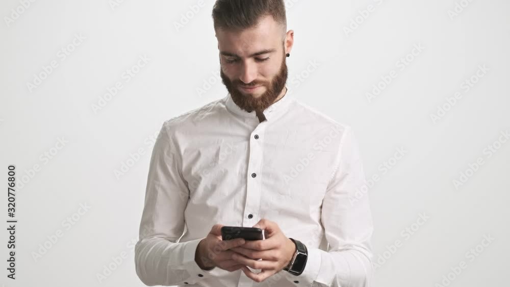 A positive calm young bearded man wearing white shirt is using a smartphone isolated over white wall background A positive calm young bearded man wearing white shirt is using a smartphone isolated over white wall background