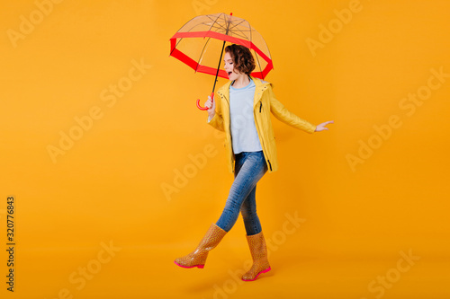 Glad curly girl in jeans funny dancing holding trendy parasol. Studio portrait of inspired young woman in rubber shoes fooling around on bright yellow background and smiling.
