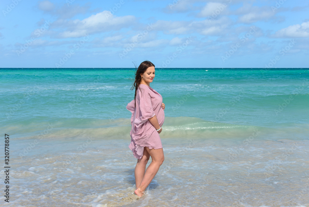 Pregnant caucasian brunette woman stands on the shore of the Atlantic ocean at midday, Cuba