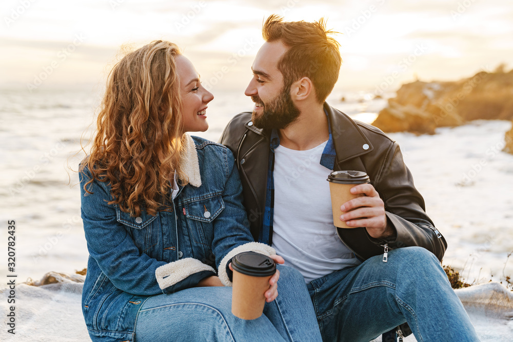 Image of beautiful young couple drinking coffee while walking by seaside
