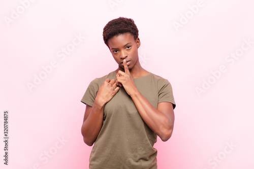 young pretty black womanlooking serious and cross with finger pressed to lips demanding silence or quiet, keeping a secret against pink wall photo