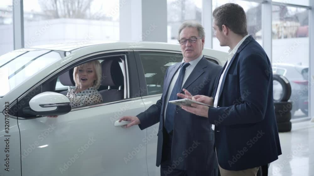 Beautiful mature Caucasian blond woman sitting in car as her senior husband talking with car dealer in showroom. Successful elderly businessman buying new vehicle for his spouse. Beautiful mature Caucasian blond woman sitting in car as her senior husband talking with car dealer in showroom. Successful elderly businessman buying new vehicle for his spouse.