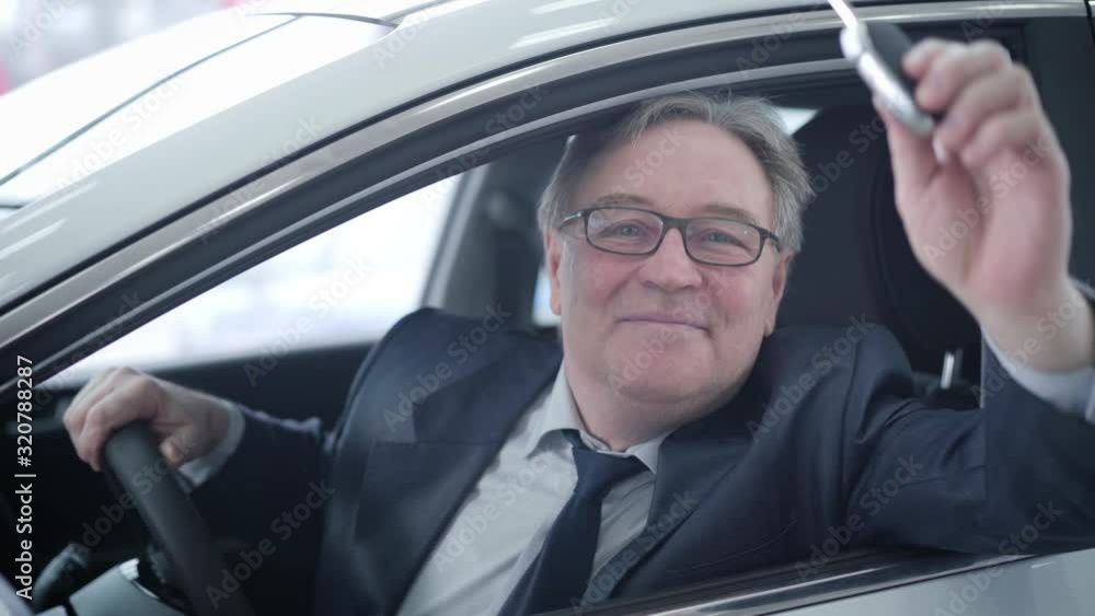 Close-up portrait of positive mature Caucasian man looking out the car window and bragging keys. Smiling senior man purchasing vehicle in car dealership. Success, automobile industry, auto showroom. Close-up portrait of positive mature Caucasian man looking out the car window and bragging keys. Smiling senior man purchasing vehicle in car dealership. Success, automobile industry, auto showroom.