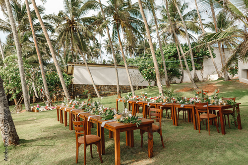 Served tables for wedding banquet with plates placed on palm leaves in restaurant. Served table for a wedding dinner in boho chic style. Decor from fresh flowers of greenery  pineapples and coconuts.