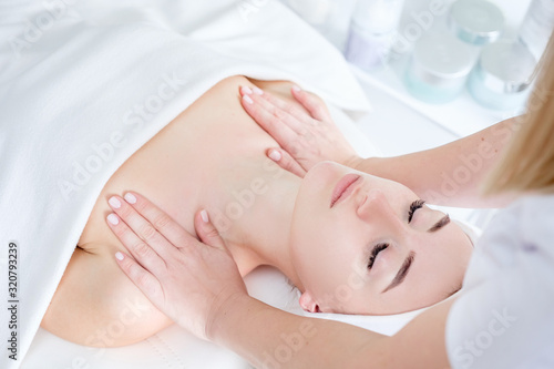 Young woman beautician doing neck and face massage to beautiful and charming young caucasian female client lying on couch under white blanket. Concept of cosmetic procedures and anti-aging products