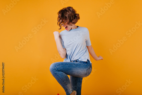 Refined short-haired lady wears dark-blue jeans jumping in studio. Attractive girl with wavy hairstyle dancing on yellow background.