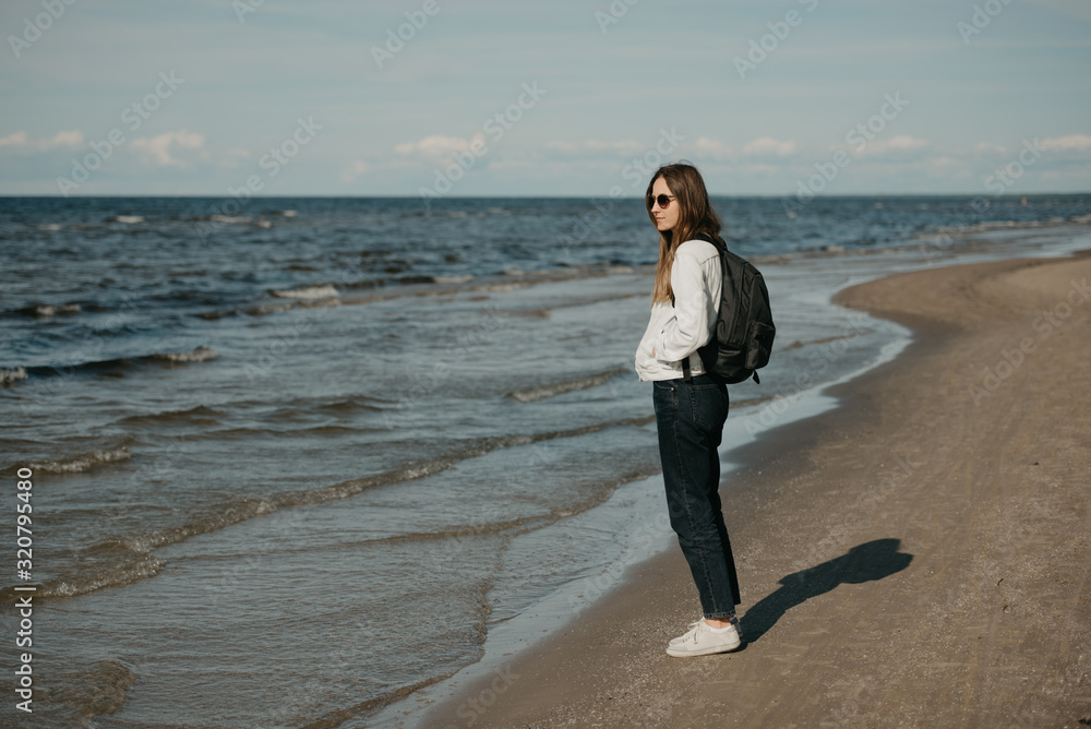 A young female tourist in a white jeans jacket, black jeans, grey sneakers, sunglasses, and backpack enjoys the sea