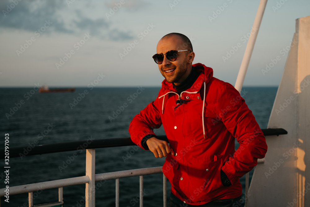 Bald brutal man with stubble in a red jacket in the sunglasses smiles aboard a ship looking at the sunset. Dry cargo ship in the background.
