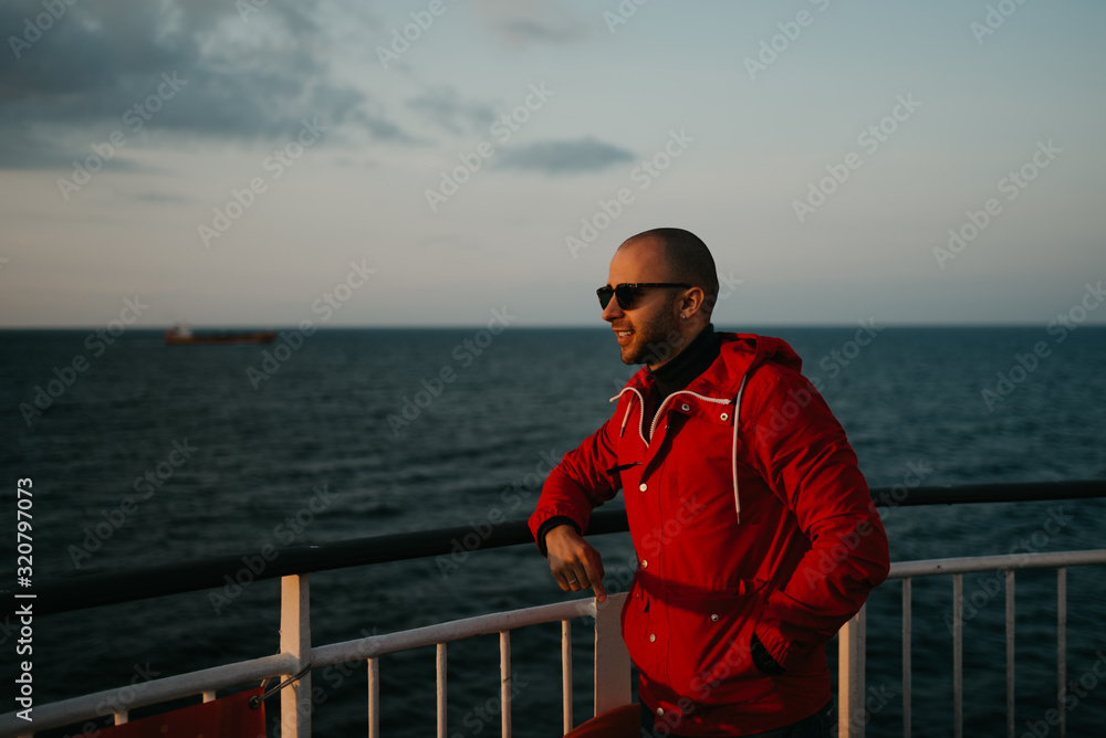Bald brutal man with stubble in a red jacket in the sunglasses with smile poses aboard a ship looking at the sunset. Dry cargo ship in the background.