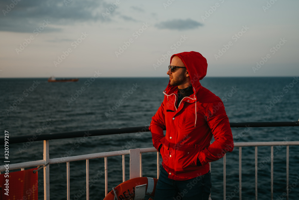A stylish man with stubble in a red jacket in the hood and in the sunglasses puts on a hood on his head aboard a ship looking at the sunset. Dry cargo ship in the background.