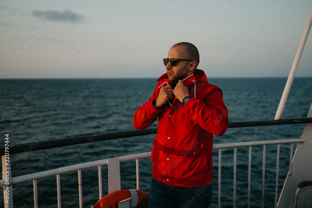 Obraz premium A bald man with stubble in a red jacket in the sunglasses fastens jacket aboard a ship looking at the sunset.