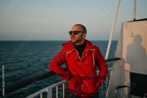 A bald man with stubble in a red jacket in the sunglasses with hands in pockets smiles aboard a ship looking at the sunset.