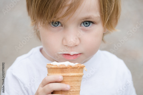 Beautiful baby with blue eyes in white tshirt is eating glass of white ice cream.