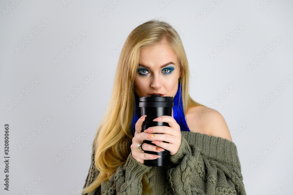 Pretty blond woman model with a cup of coffee in her hands with long hair and excellent make-up is standing in front of the camera on a white background in a green jacket. Shows different emotions.
