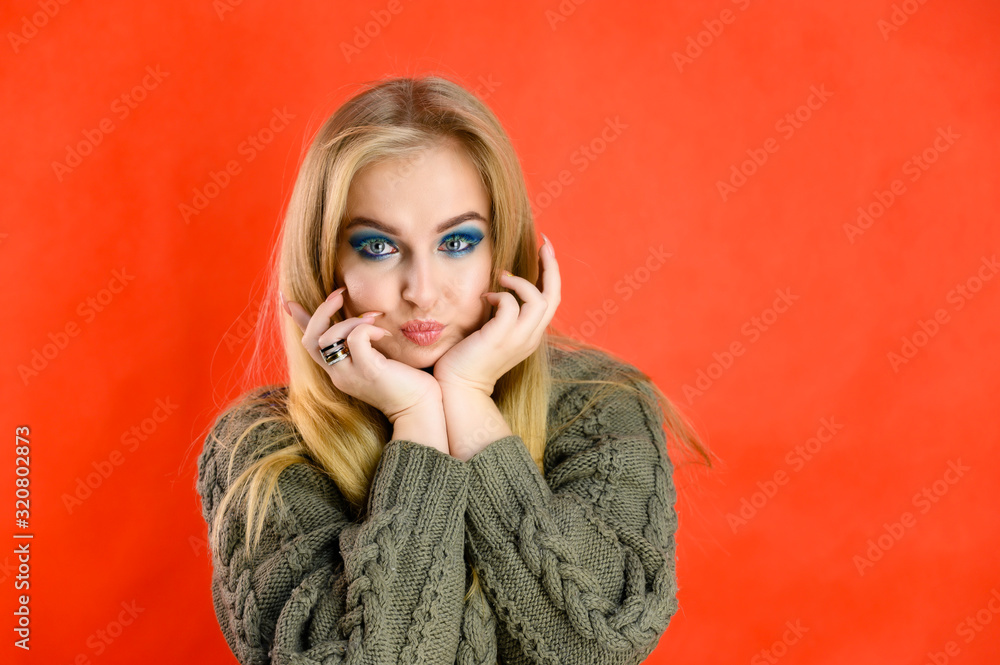 Nice girl model blonde with long hair and excellent make-up is standing in front of the camera on a red background in a green sweater. A woman shows different emotions. Nice girl model blonde with long hair and excellent make-up is standing in front of the camera on a red background in a green sweater. A woman shows different emotions.