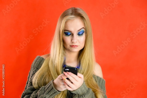 A nice girl with a phone in her hands with long hair and excellent make-up is standing in front of the camera on a red background in a green jacket. A woman shows different emotions.