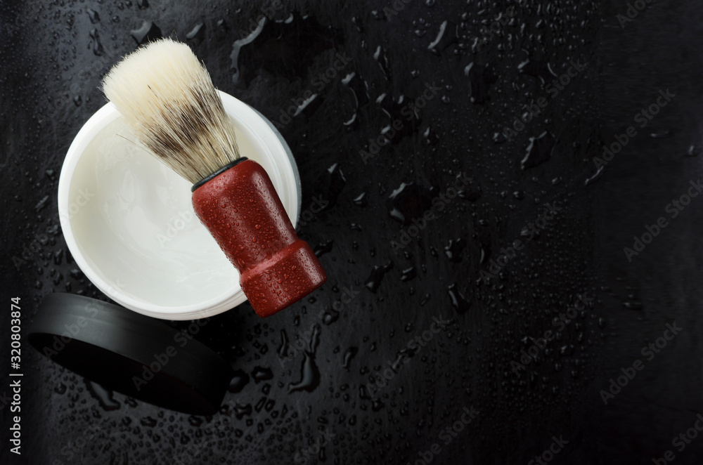 Top view of jar of foam for shaving and brush with wooden handle, black surface with water drops.Empty space for text Top view of jar of foam for shaving and brush with wooden handle, black surface with water drops.Empty space for text