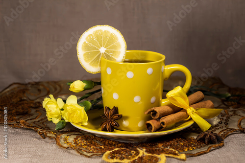A cup of tea with lemon is on a napkin. On a saucer lies star anise, cinnamon sticks and yellow clove flowers.