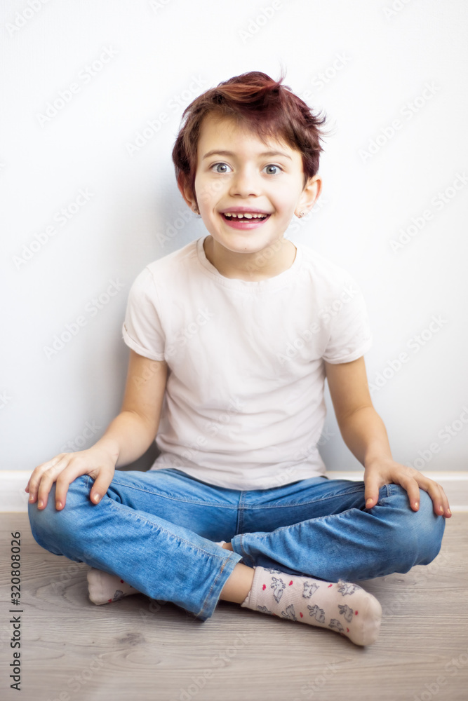 Vertical picture of smiling child with short haircut and red hair sitting on floor in blue jeans. Stylish modern happy girl with cool haircut. Happy childhood in cozy home. Copy space for text Vertical picture of smiling child with short haircut and red hair sitting on floor in blue jeans. Stylish modern happy girl with cool haircut. Happy childhood in cozy home. Copy space for text