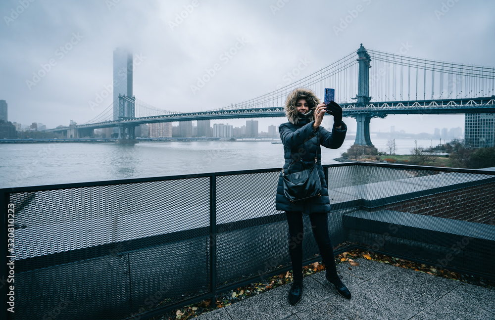 Tourist woman with warm clothes taking a selfie with the Manhattan Bridge on the background in DUMBO neighborhood while sightseeing new york during winter season. t