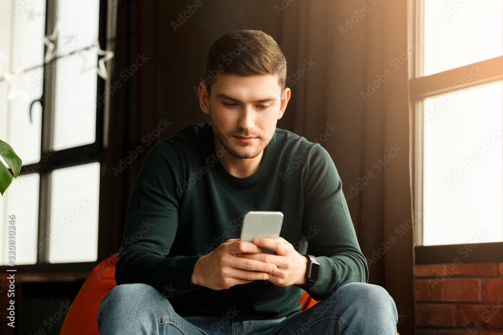 Millennial Guy Using Smartphone Texting Sitting In Beanbag Chair Indoor Millennial Guy Using Smartphone Texting Sitting In Beanbag Chair Indoor