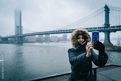 Tourist woman with warm clothes taking a selfie with the Manhattan Bridge on the background in DUMBO neighborhood while sightseeing new york during winter season. t