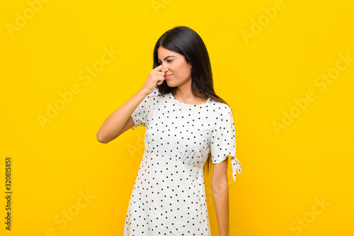 young pretty latin woman feeling disgusted, holding nose to avoid smelling a foul and unpleasant stench against flat wall