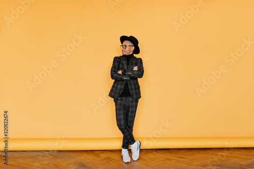 Full-length photo of stylish african man in white shoes posing in studio. Portrait of handsome black male model standing with legs crossed on yellow background.